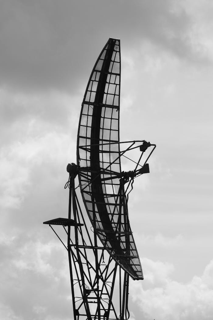 Services-02 Black and white image of a large radar antenna structure against a cloudy sky, emphasizing technology and communication.